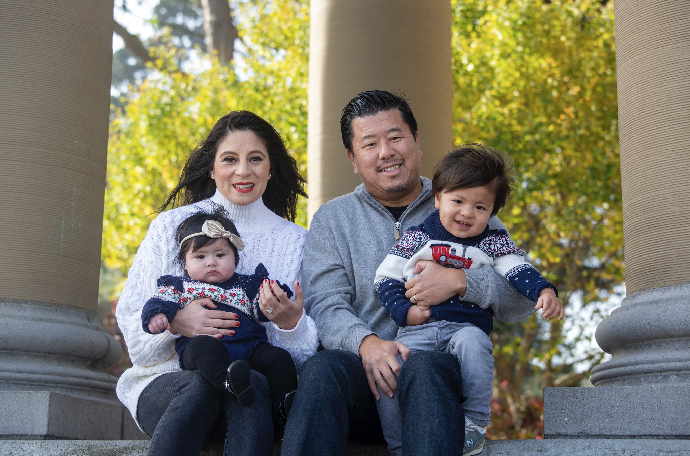 Family of four posing outdoors, seated on steps with a backdrop of trees and columns.