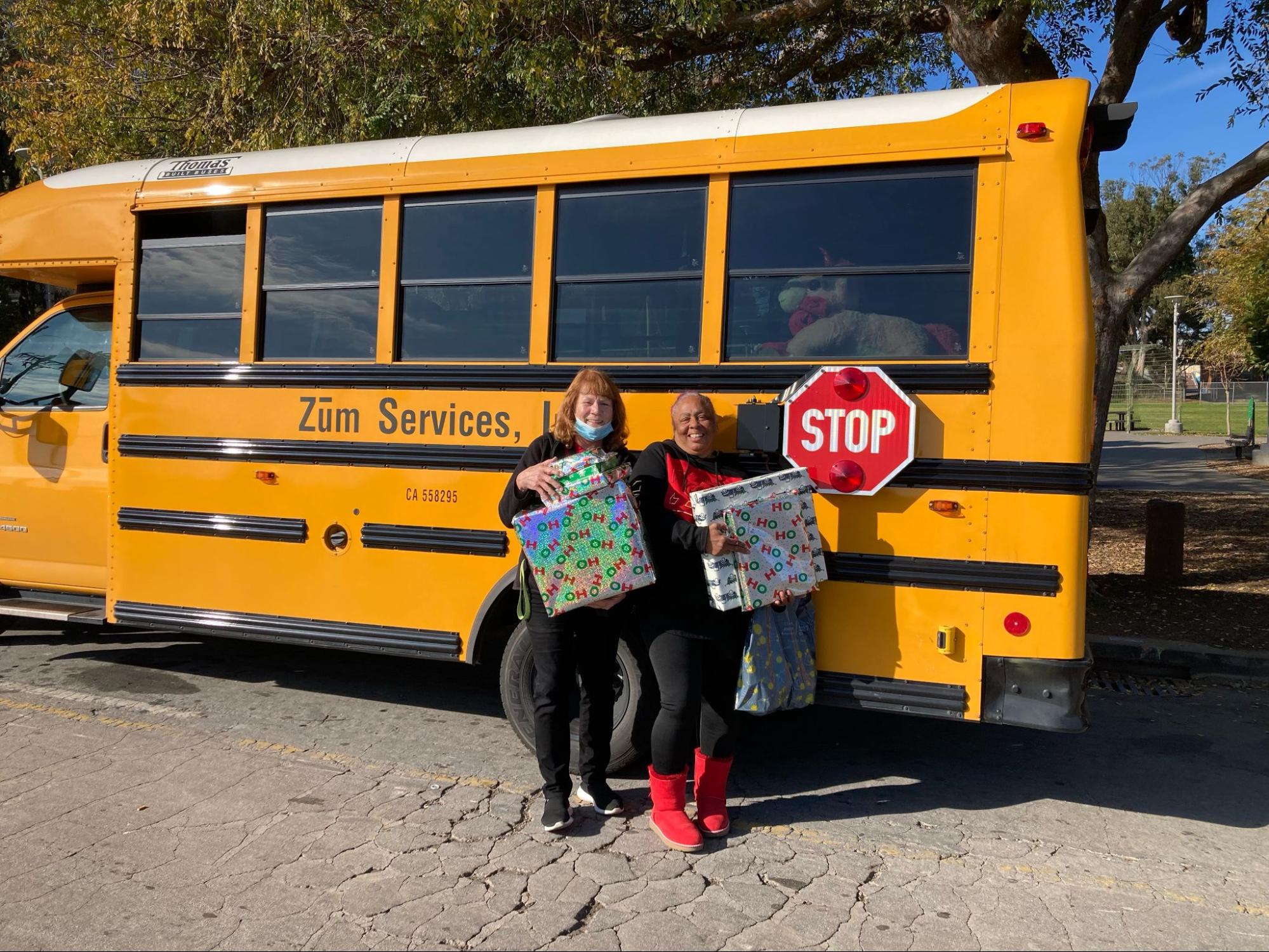 Two Zum bus drivers stand by a yellow bus, holding wrapped gifts for families in need.
