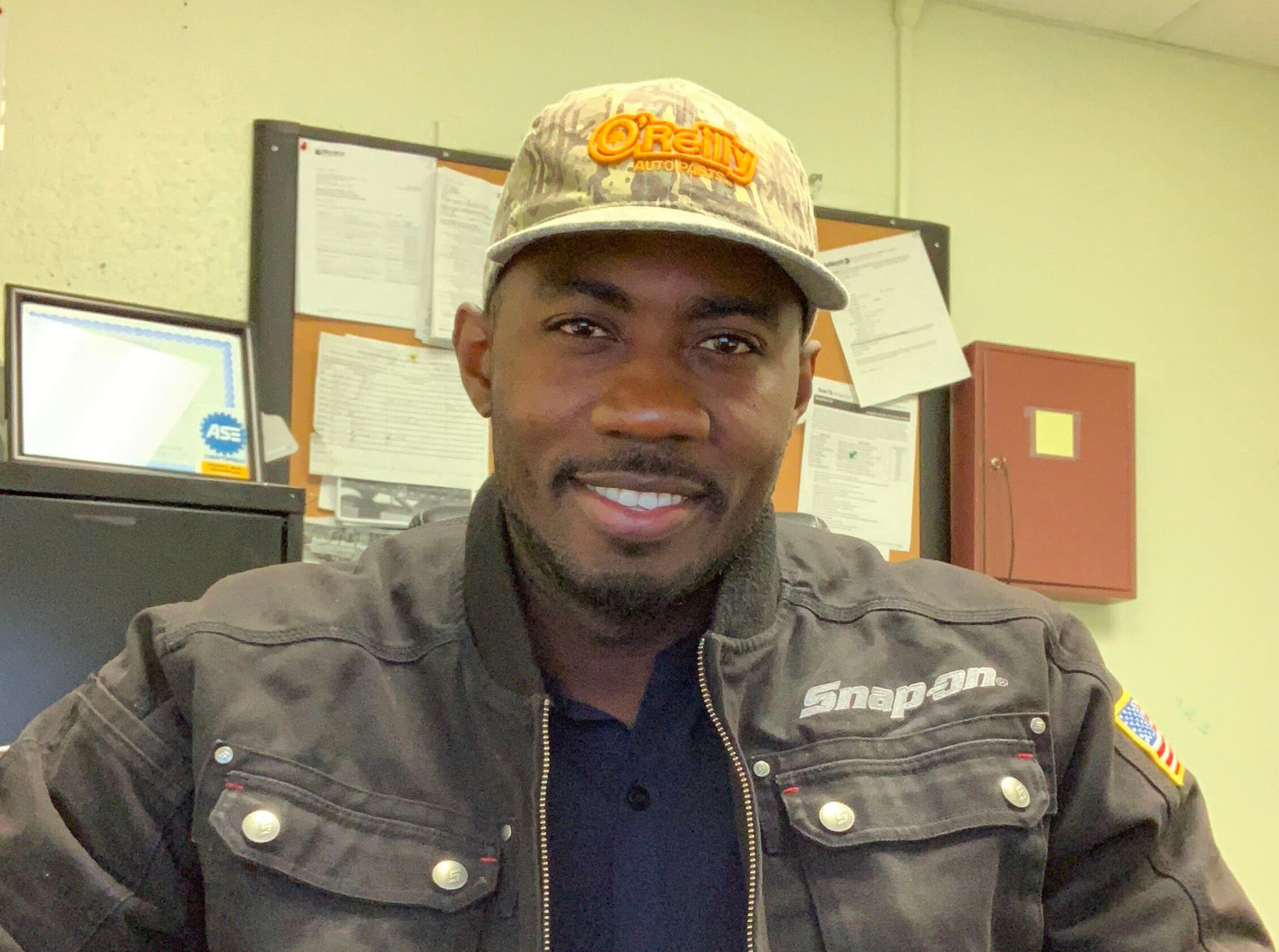 A man wearing a Snap-on jacket and O'Reilly cap sits at a desk smiling.