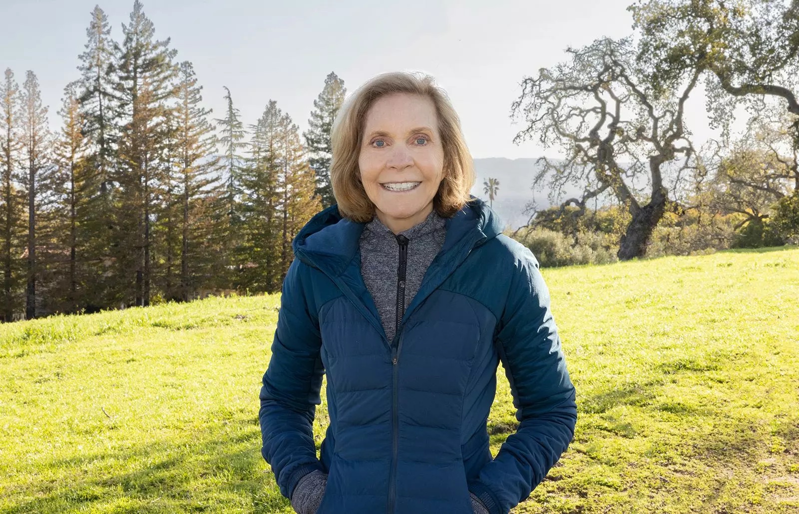 Lynn Miller smiles while standing on green grass with trees in the background.
