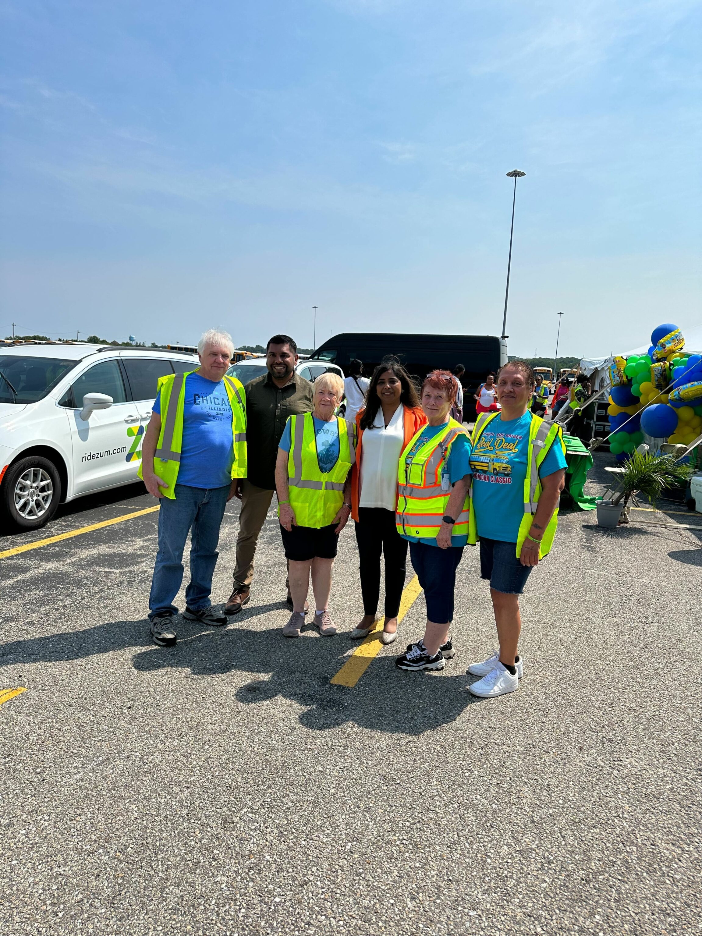Six individuals in safety vests pose together at a community event for Zum in a parking lot.