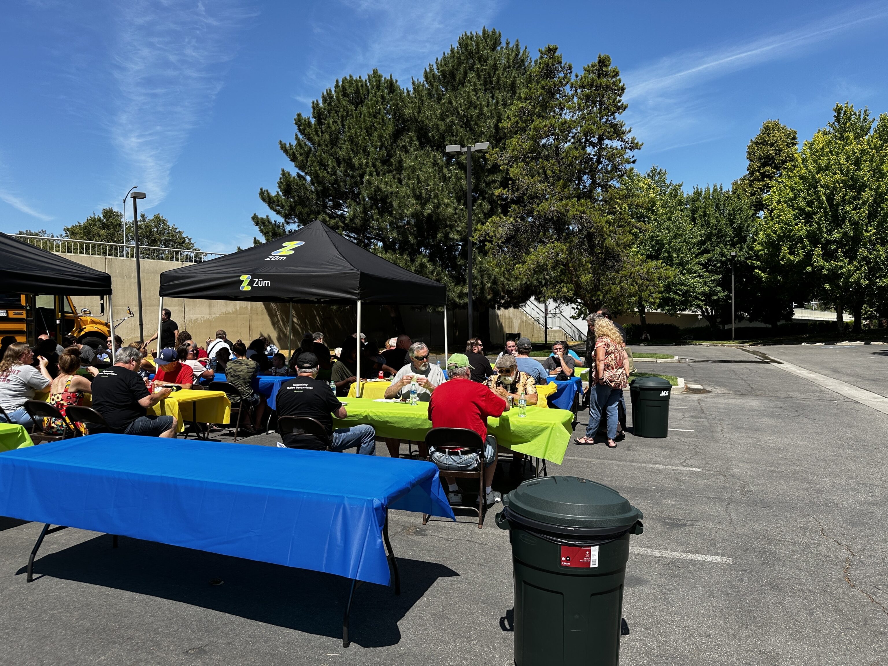 Employees gather around tables under tents, enjoying a meal in a parking lot with trees in the