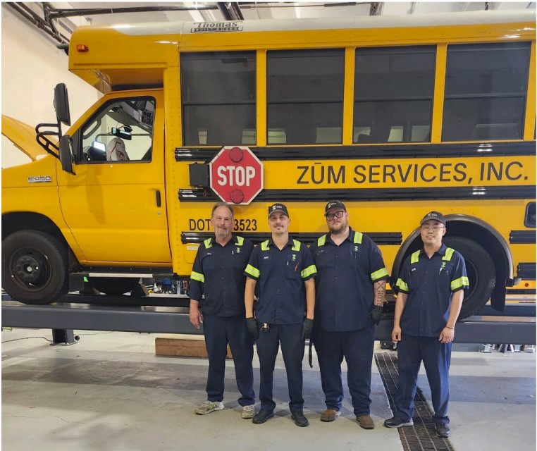 Four Zum Services employees stand in front of a yellow school bus in a garage setting.
