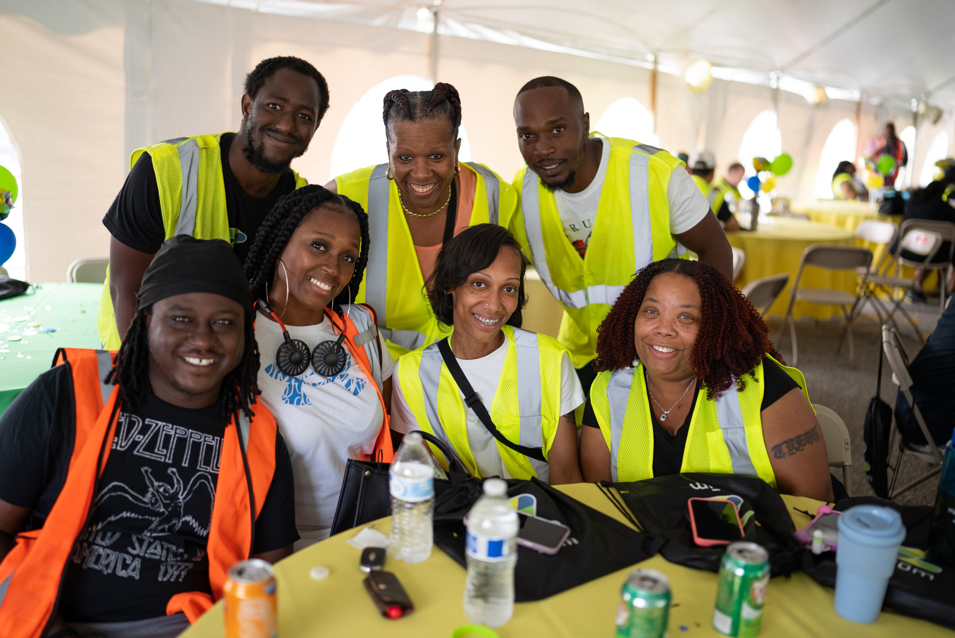 Seven people in yellow safety vests pose together at a table with drinks during an event.