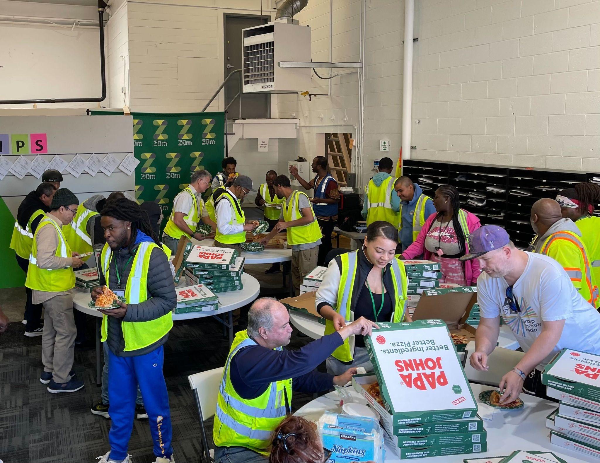 Volunteers in yellow vests assemble pizza boxes in a workspace, promoting teamwork and community.
