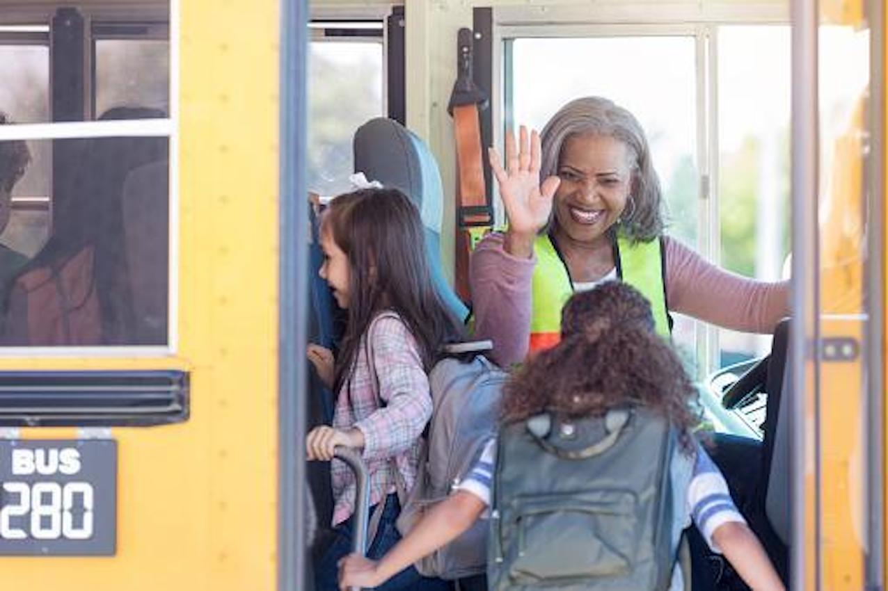 A bus driver waves as students board a school bus, with smiling faces and backpacks.