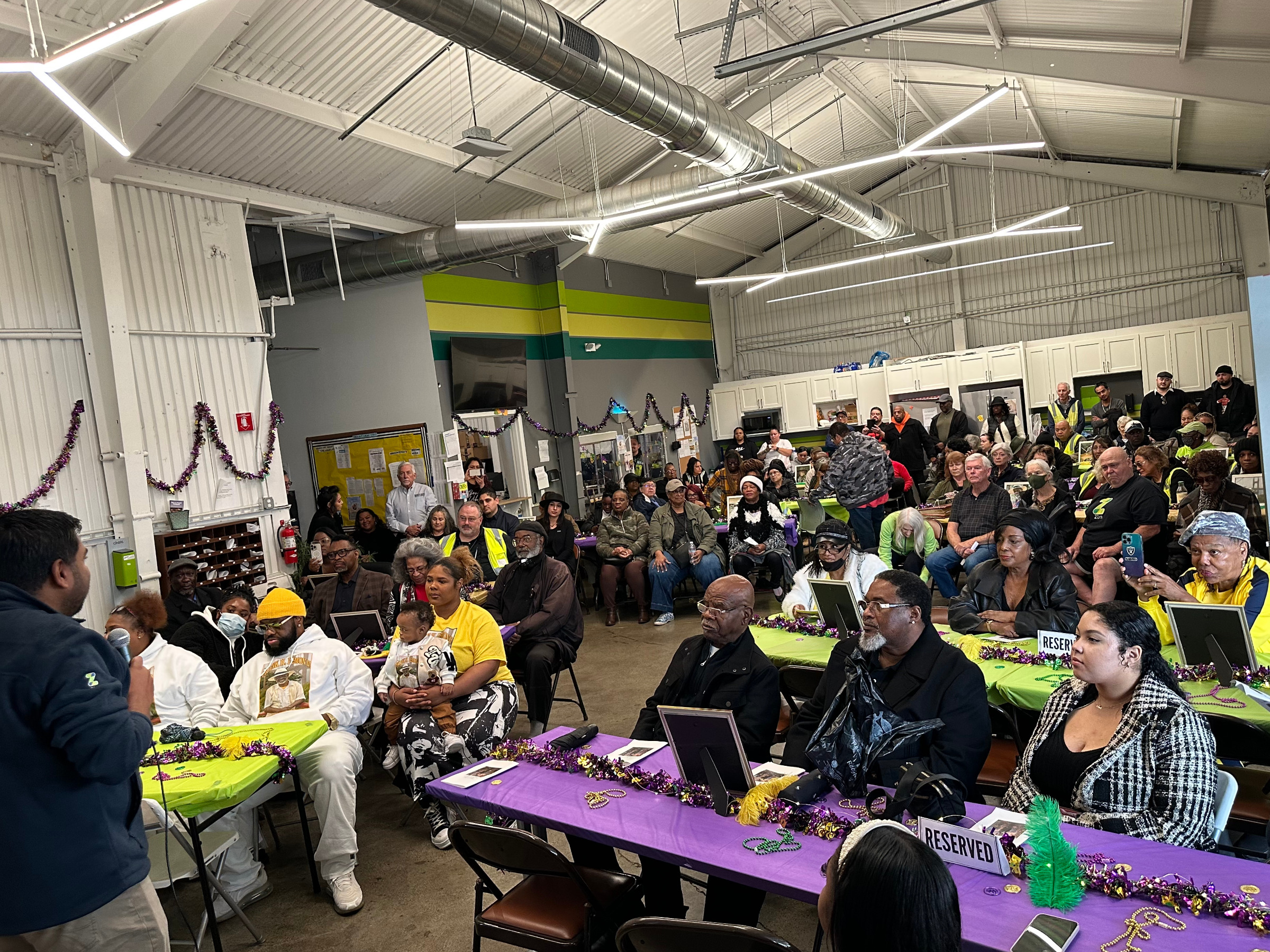 A large group gathers indoors at an event, celebrating with decorations and a speaker at the front.