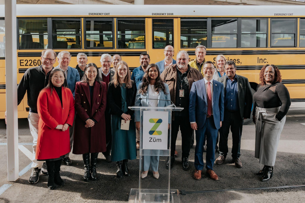 Group of Zum representatives and officials gathered in front of yellow school buses at an event.