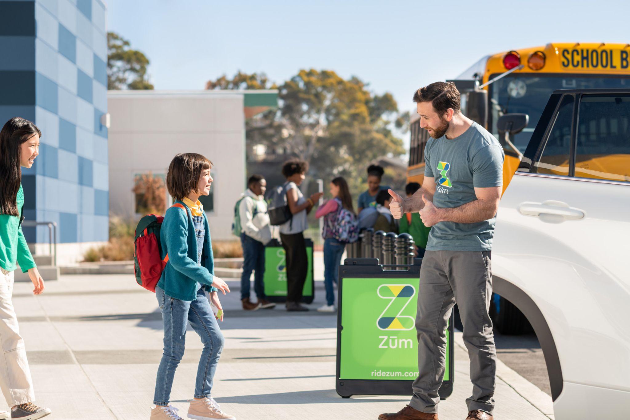 A driver greets a student by a Zum vehicle as others wait nearby for school transportation.