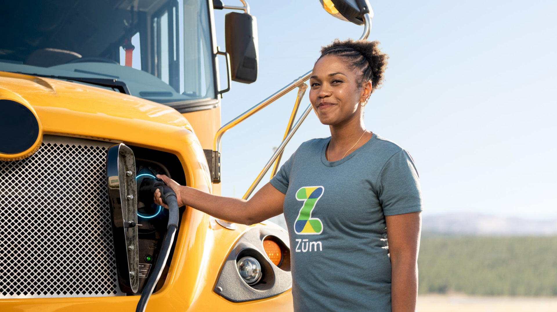 A woman in a Zum. t-shirt charges an electric bus, smiling near the vehicle in a sunny environment.