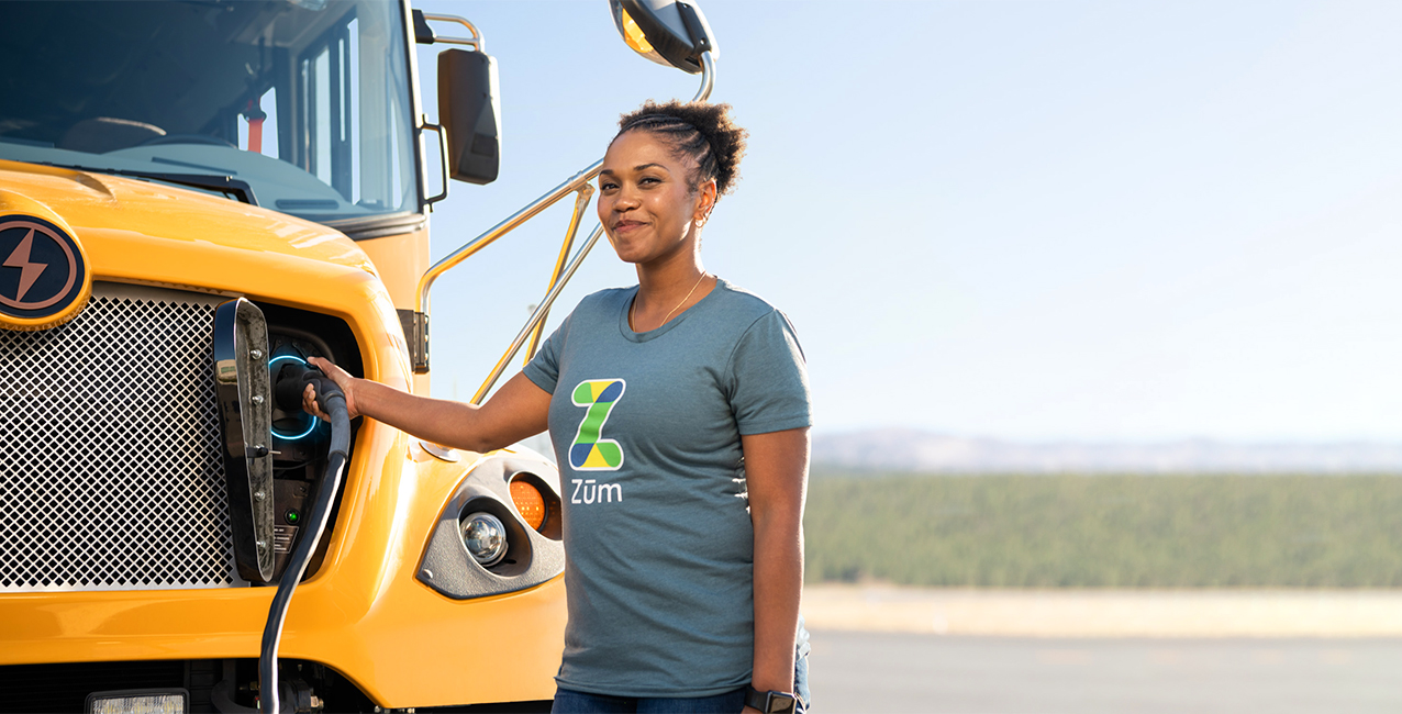 A woman in a Zum t-shirt connects an electric bus to a charging station outdoors.