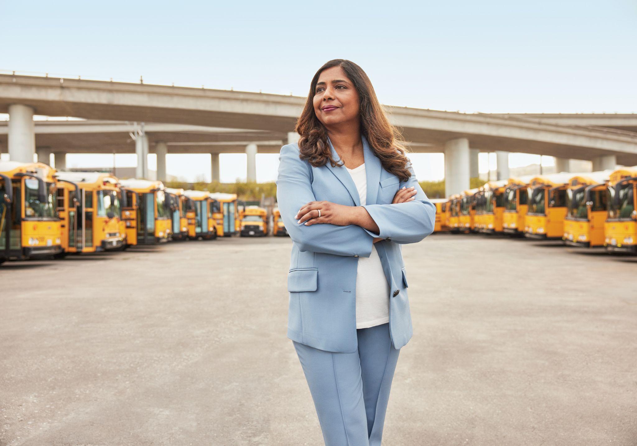 A woman in a light blue suit stands confidently among yellow school buses in a yard.