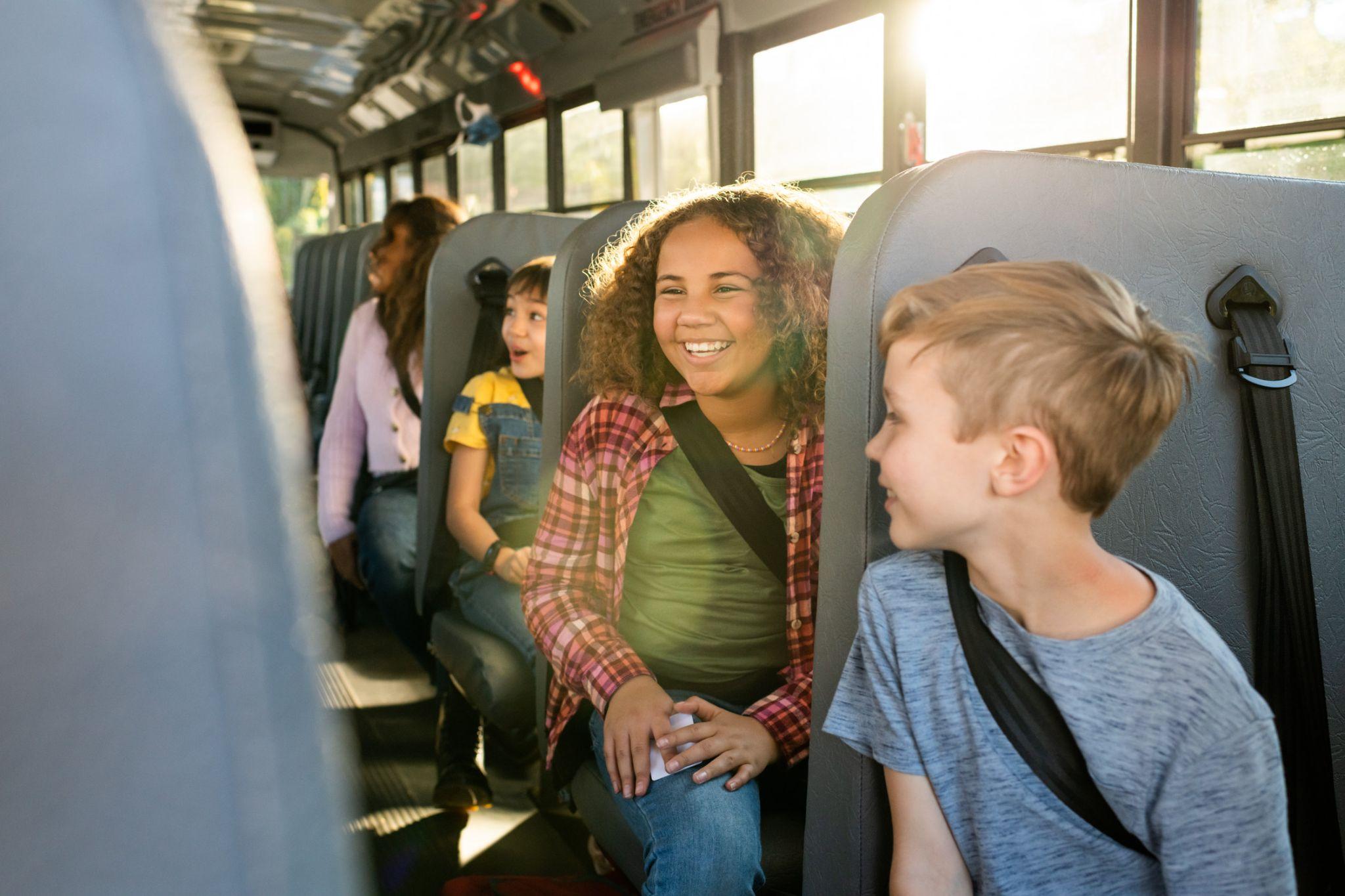 Students smiling and chatting on a school bus, with sunlight filtering through the windows.