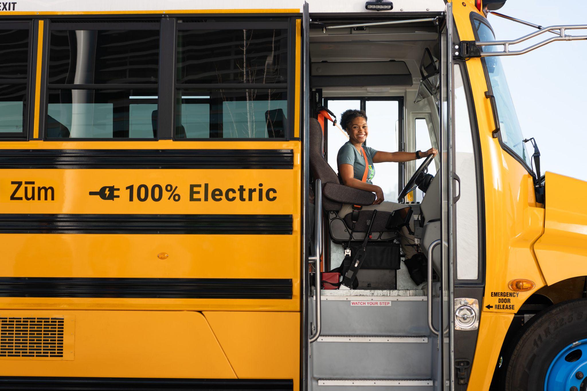 A driver inside a yellow electric school bus labeled "Zum 100% Electric."