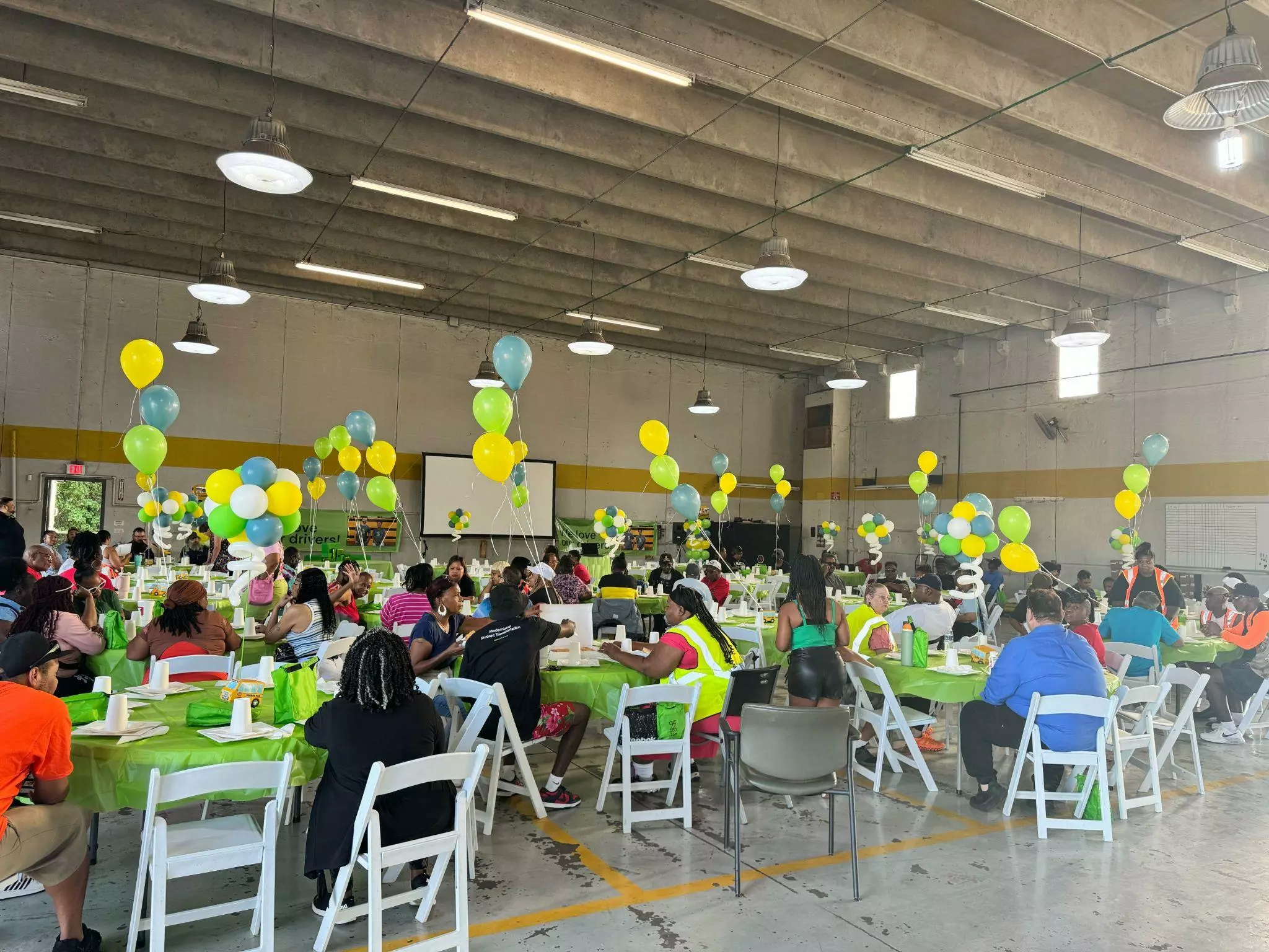 A spacious hall filled with people at tables, decorated with yellow and green balloons.