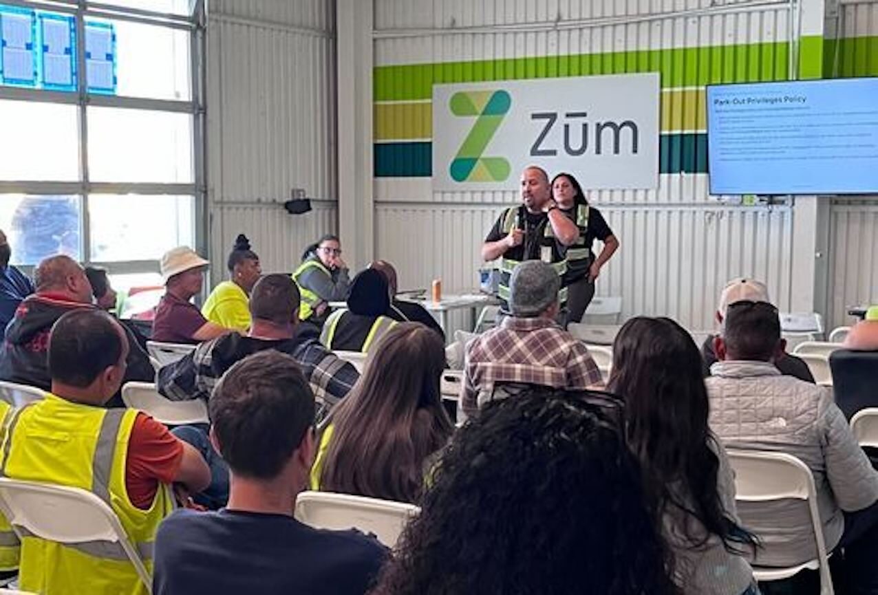 A group meeting at Zum, with staff presenting to attendees in reflective vests.