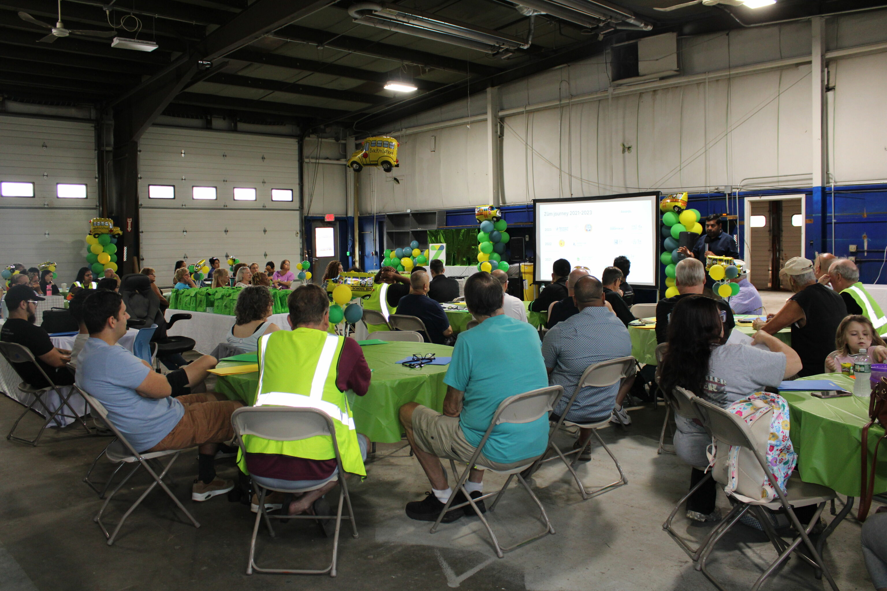 Audience gathered in a large hall with green tables and balloons for a Zum event presentation.