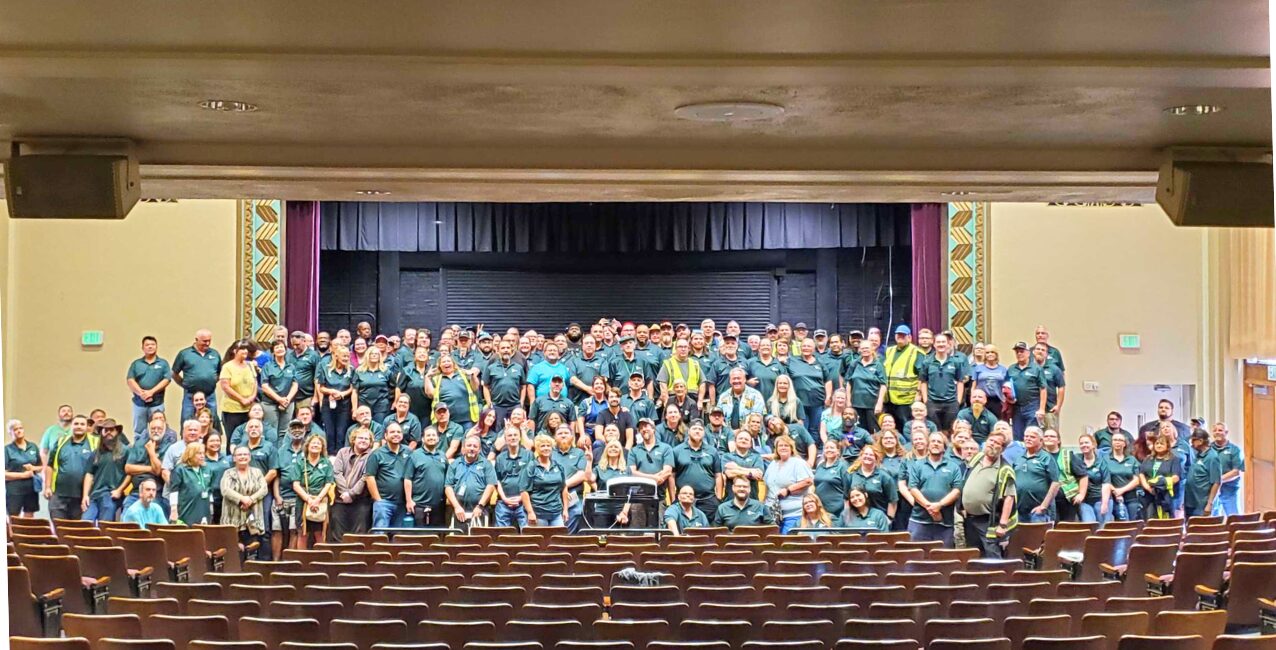 A large group of Zum employees poses together on stage in a theater, all wearing teal shirts.
