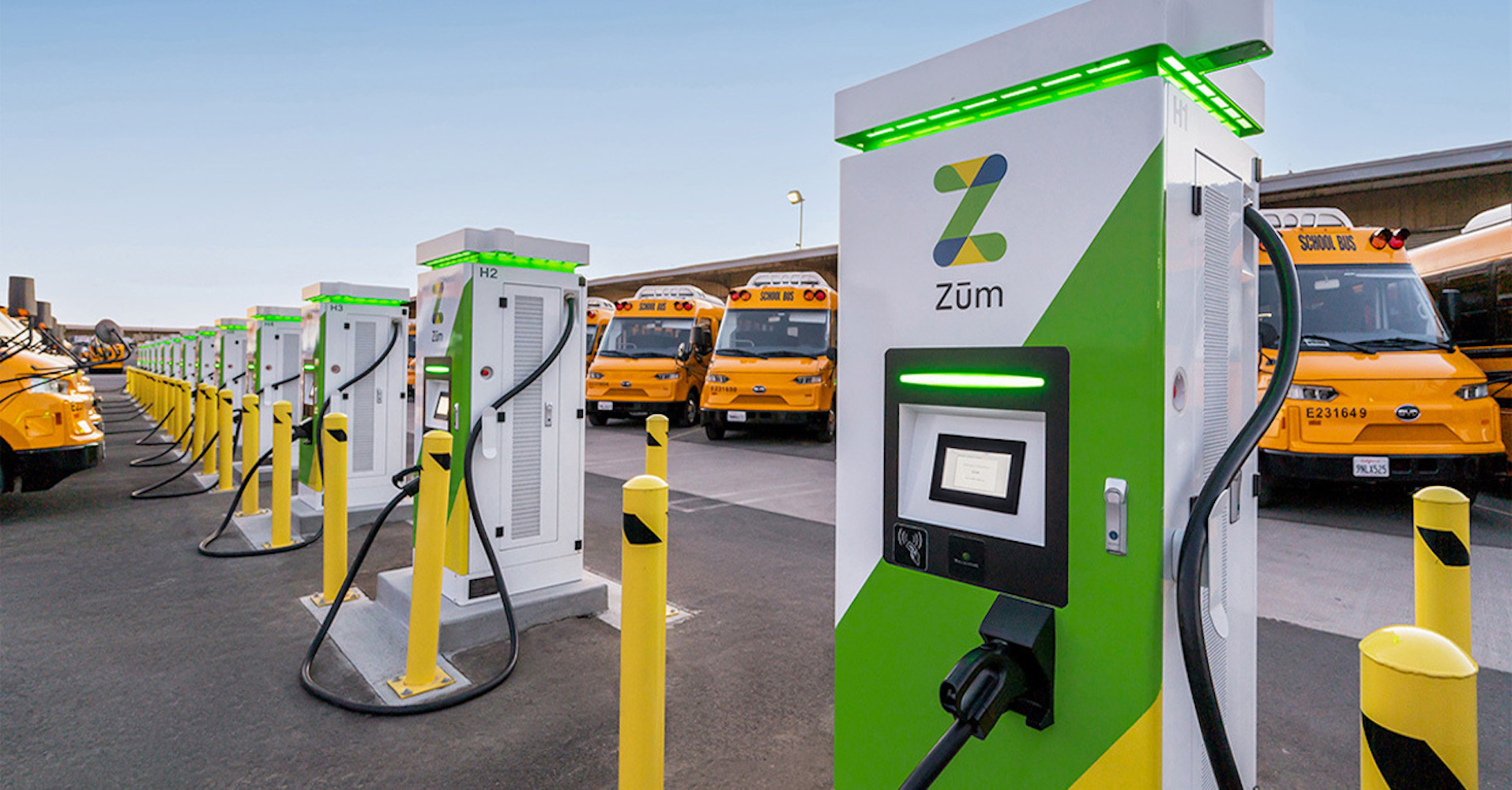 Electric vehicle chargers lined up in a bus yard, with yellow school buses in the background.