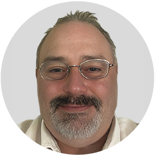 Close-up of a smiling man with glasses and a beard, wearing a light-colored shirt.