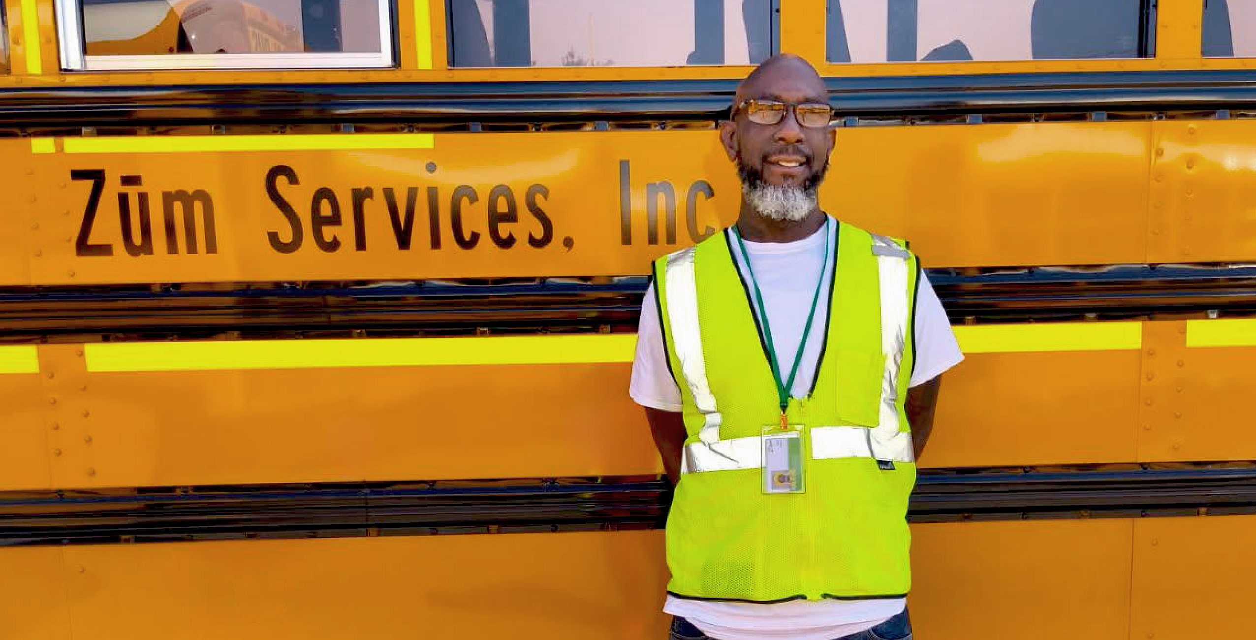 Zum driver stands in front of a yellow bus, wearing a safety vest and glasses, smiling.