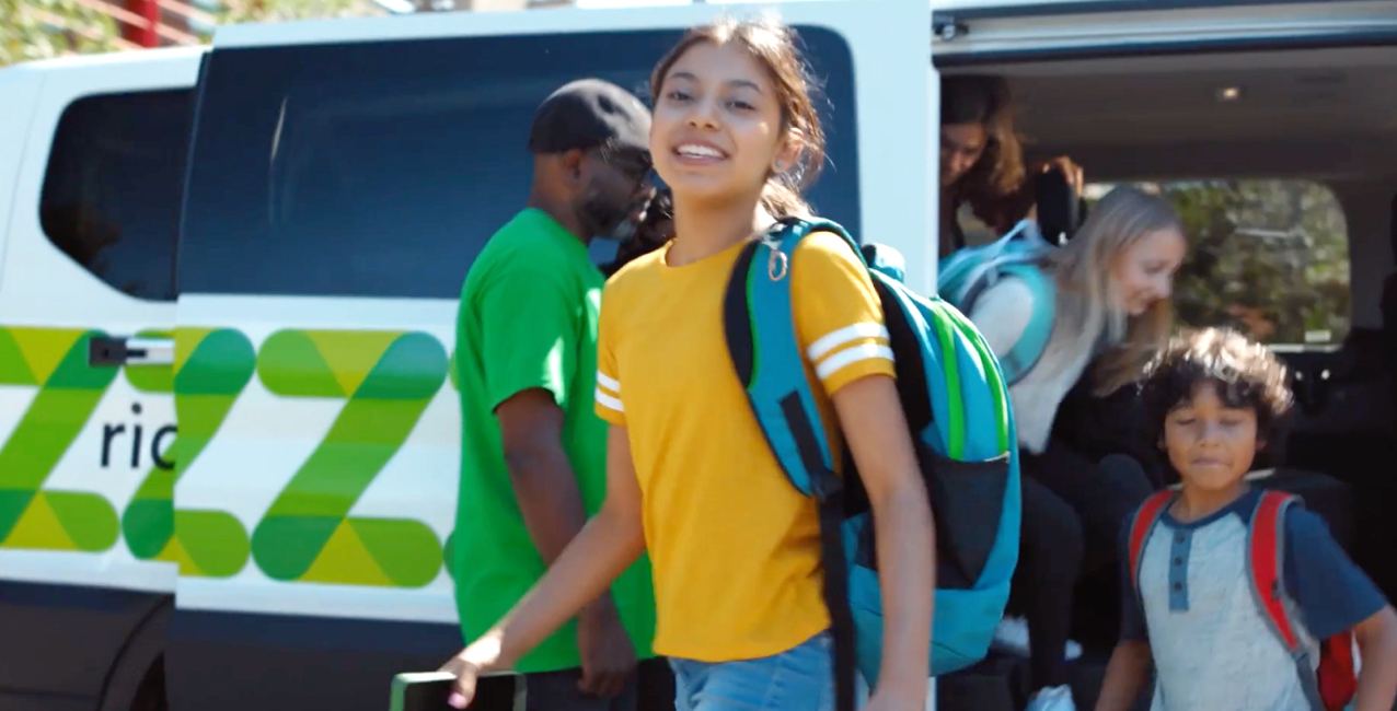 A girl in a yellow shirt smiles while walking towards a van, with children and adults nearby.