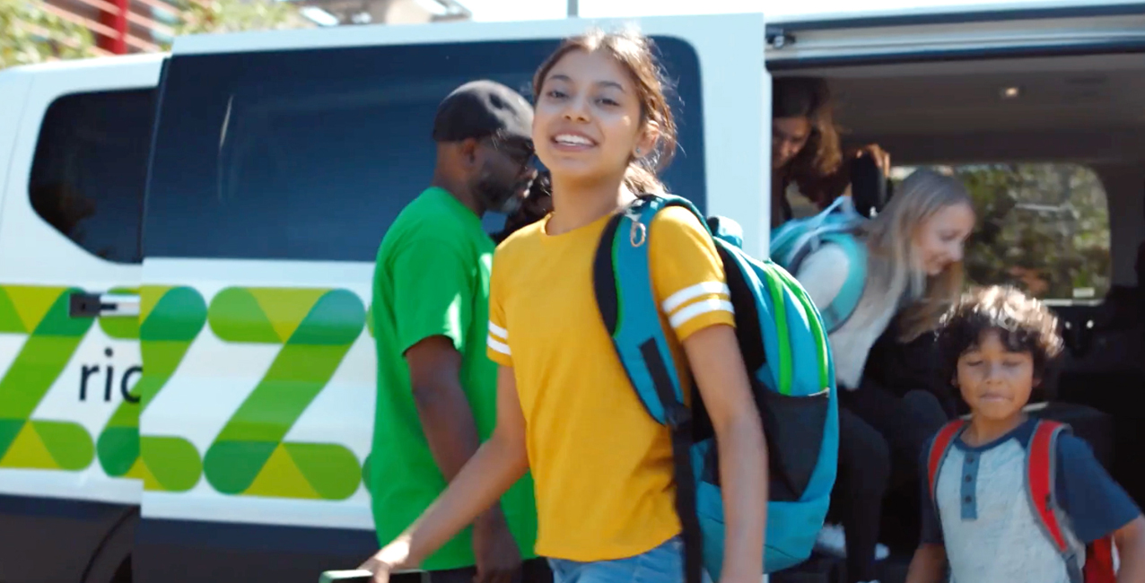 Girl smiling while walking towards a van, with other children and adults in the background.