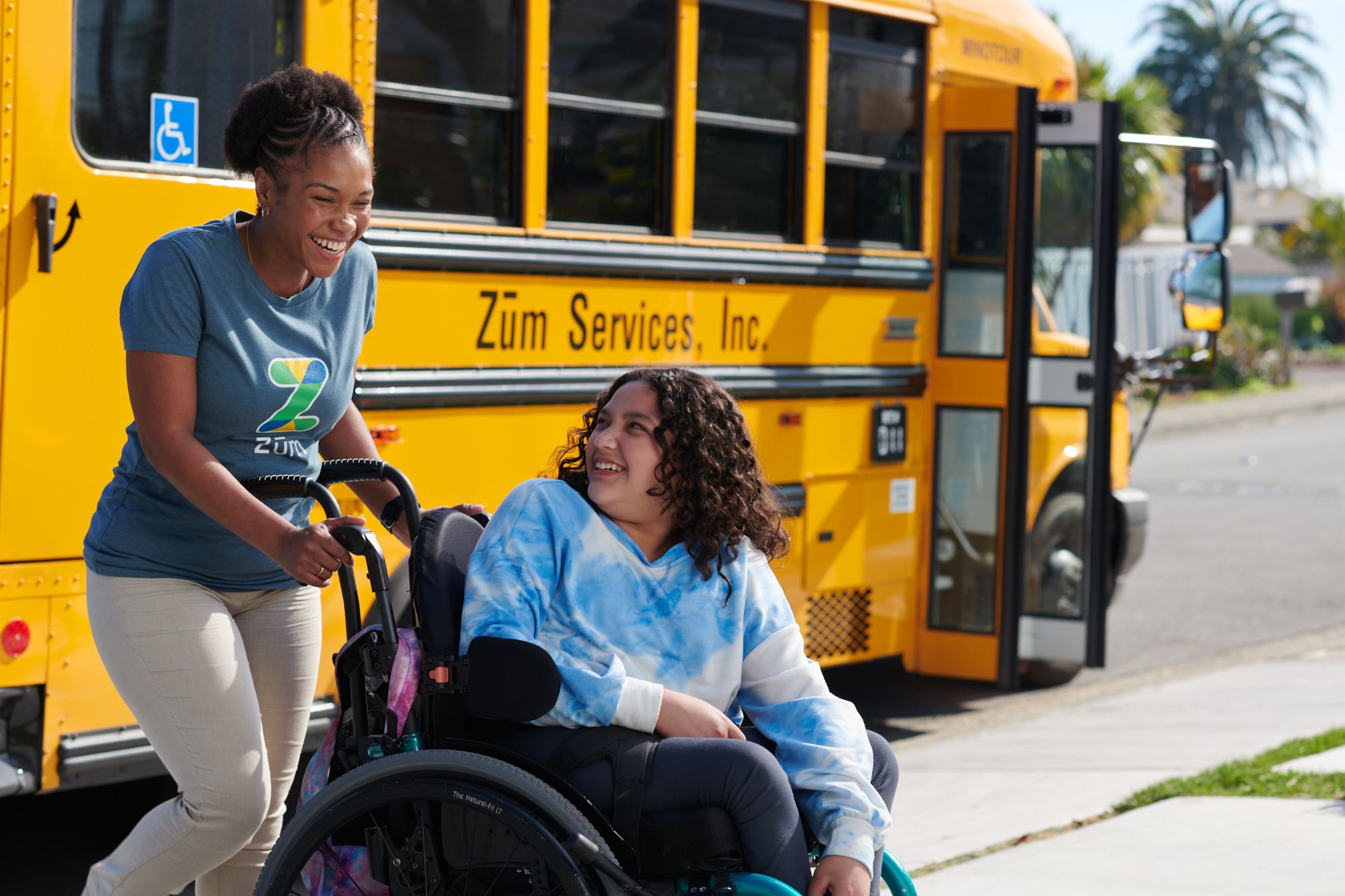 A caregiver assists a smiling student in a wheelchair near a yellow school bus.