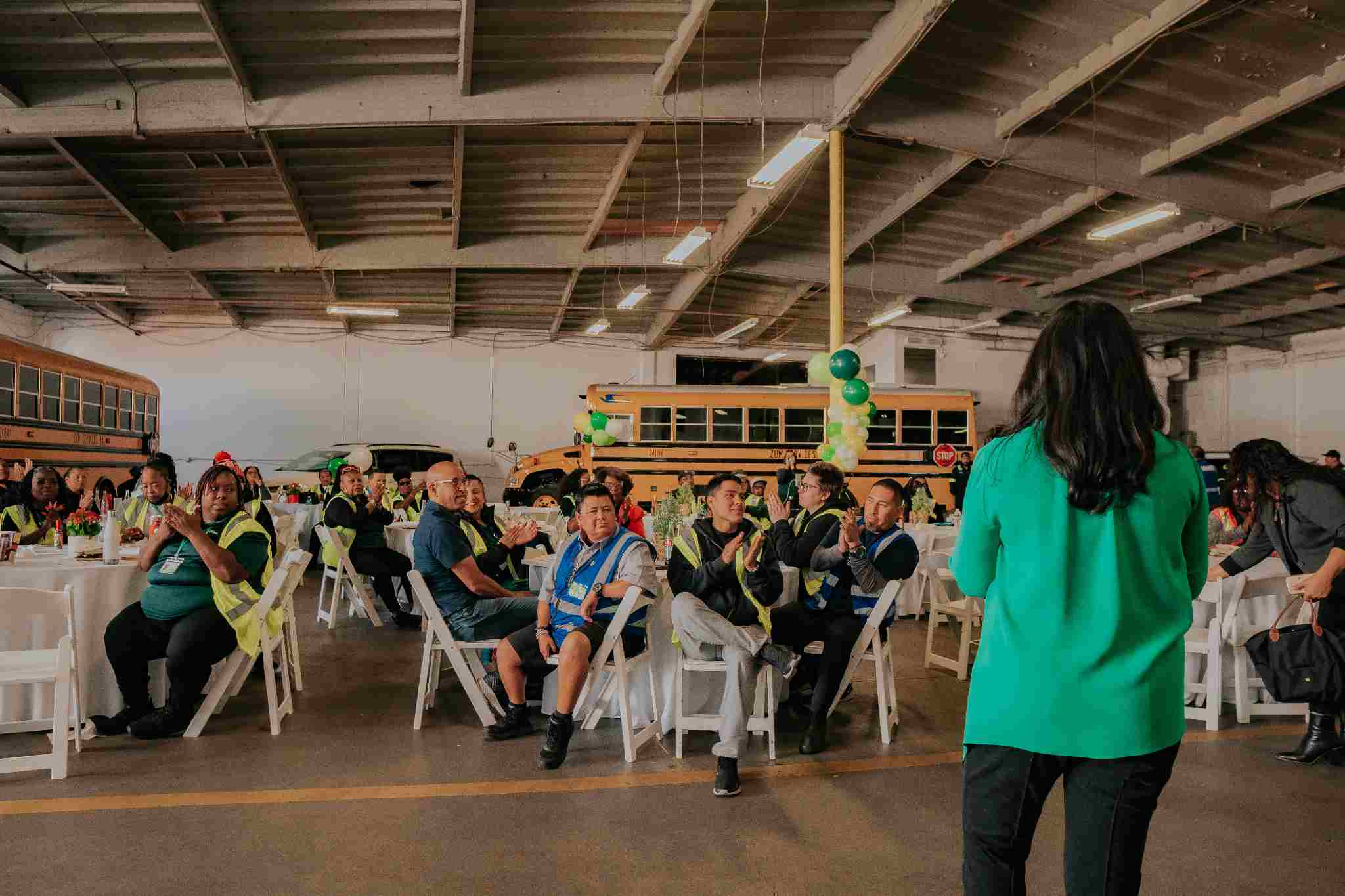 Event attendees seated at tables in a bus garage, listening to a speaker in front.