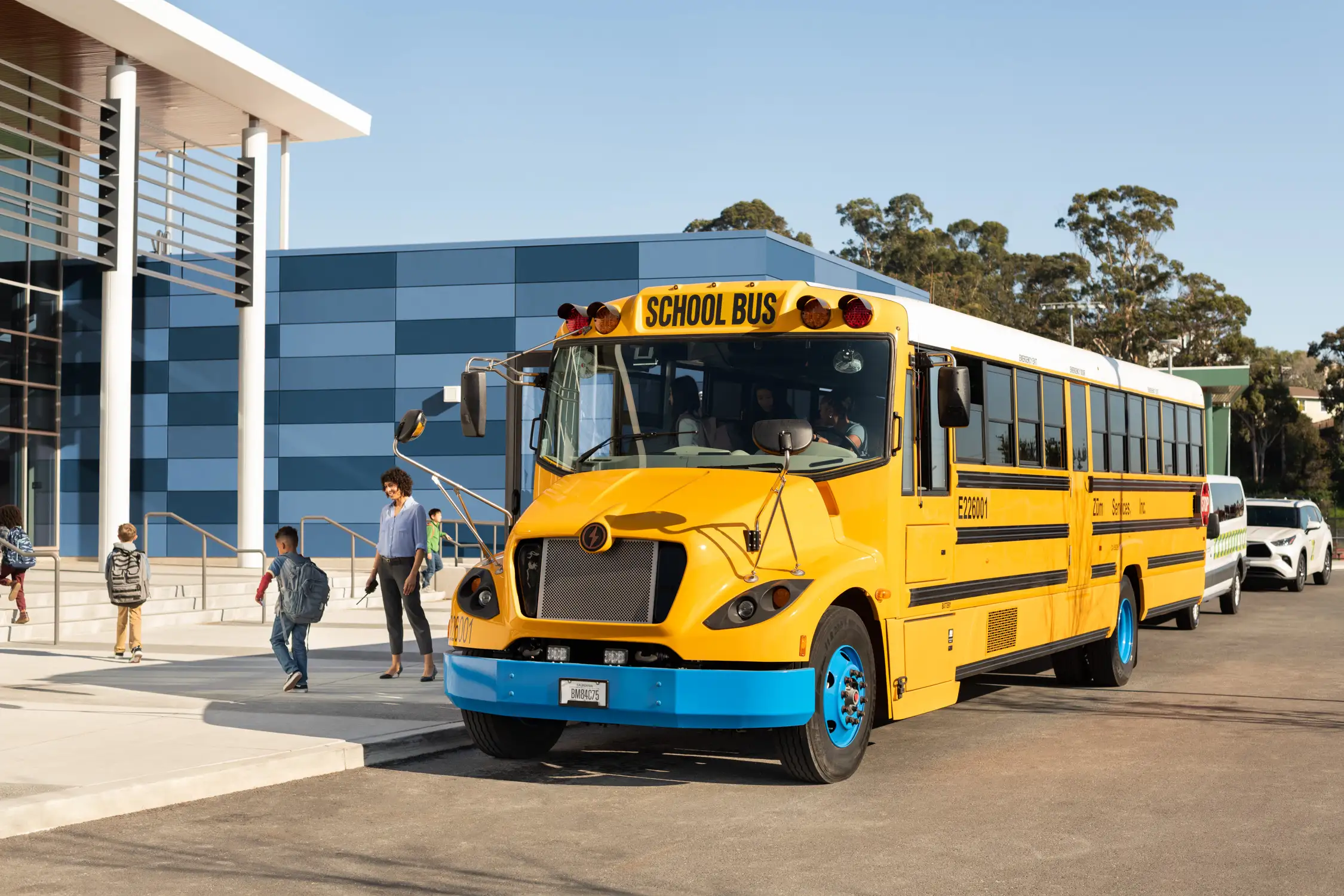 Yellow school bus parked outside a building with students walking nearby and a car in the