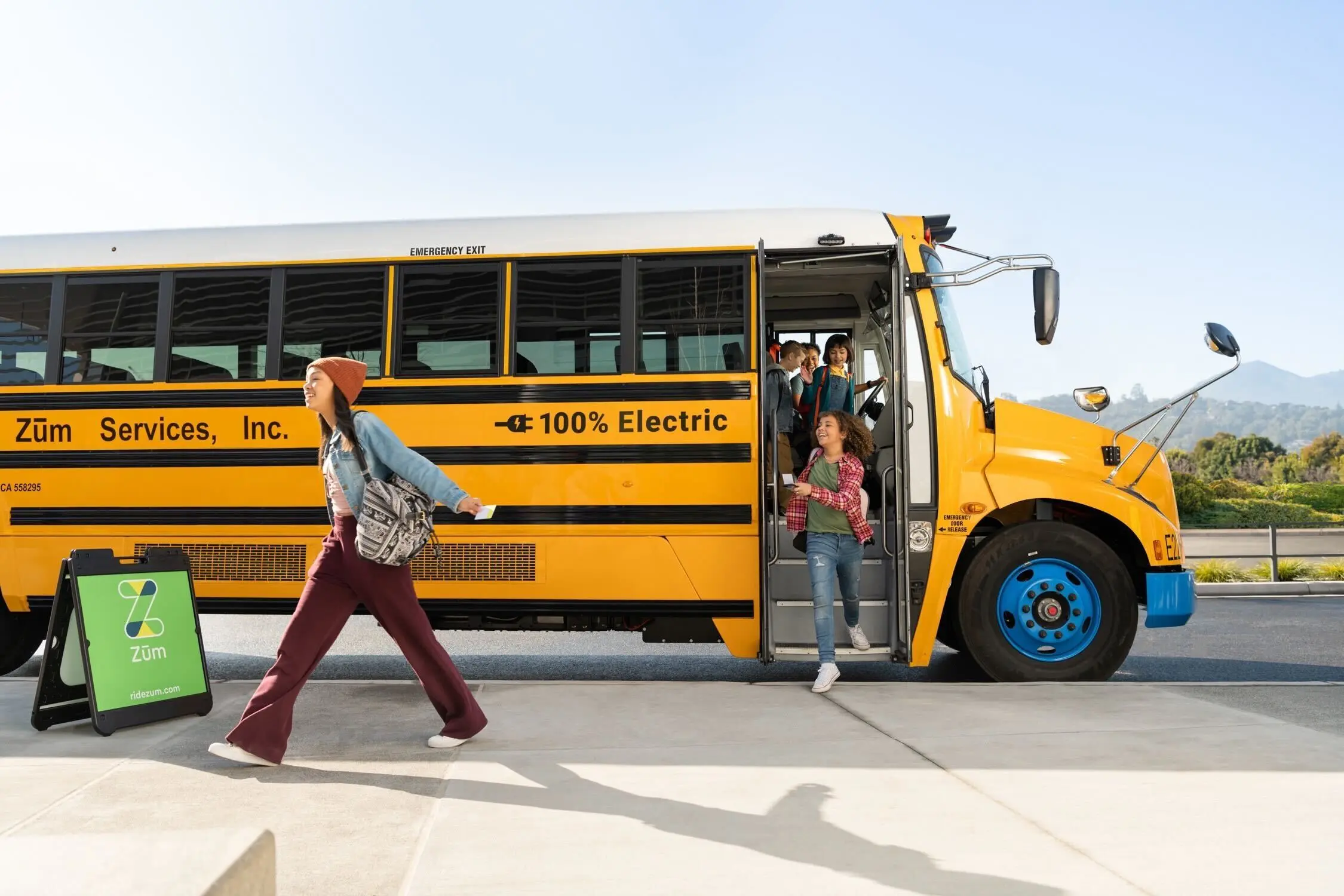Students disembark from a yellow electric bus labeled "100% Electric" with Zum Services, Inc.