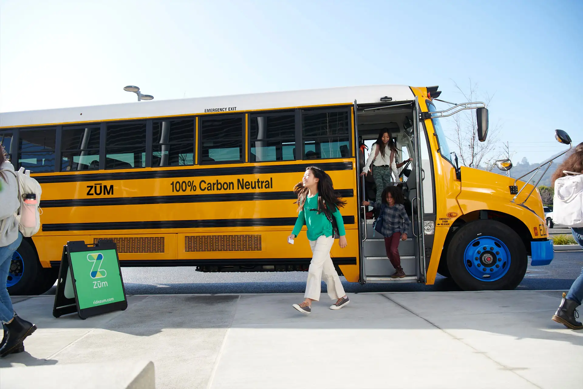 Students exit a yellow Zum bus marked "100% Carbon Neutral" in a school setting.