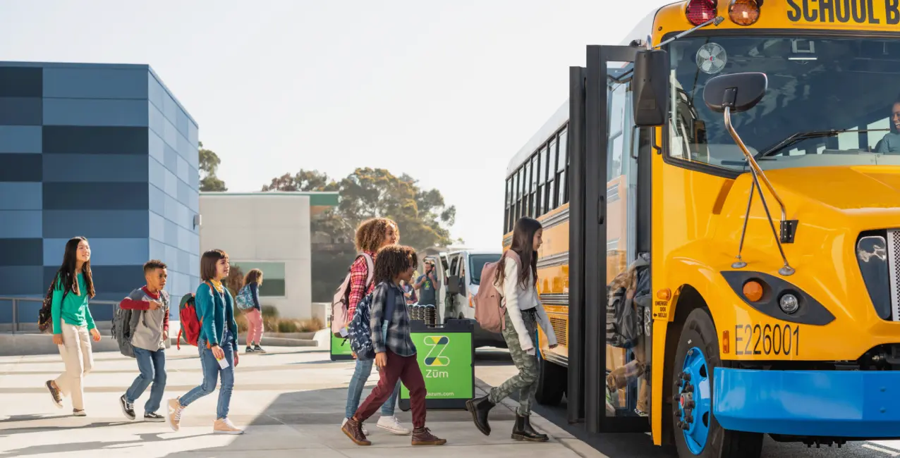 Students boarding a yellow school bus outside a modern building, with a Zum logo in view.