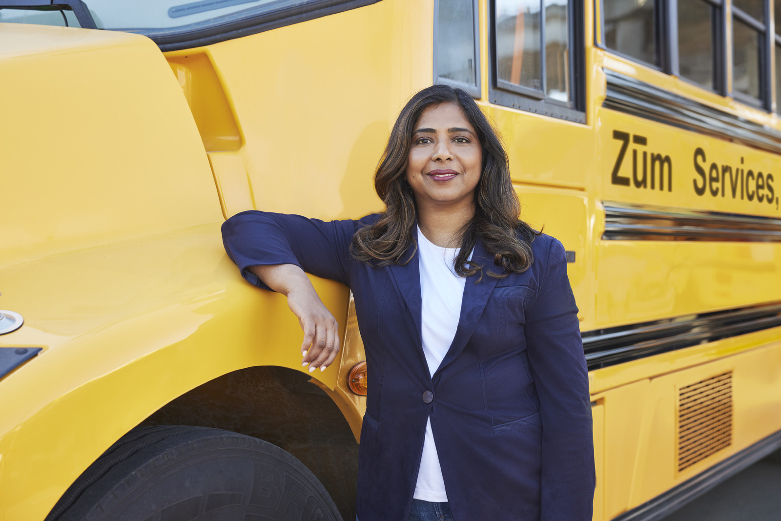 Ritu Narayan, Zum CEO, stands beside a yellow bus, smiling, in a professional outfit.