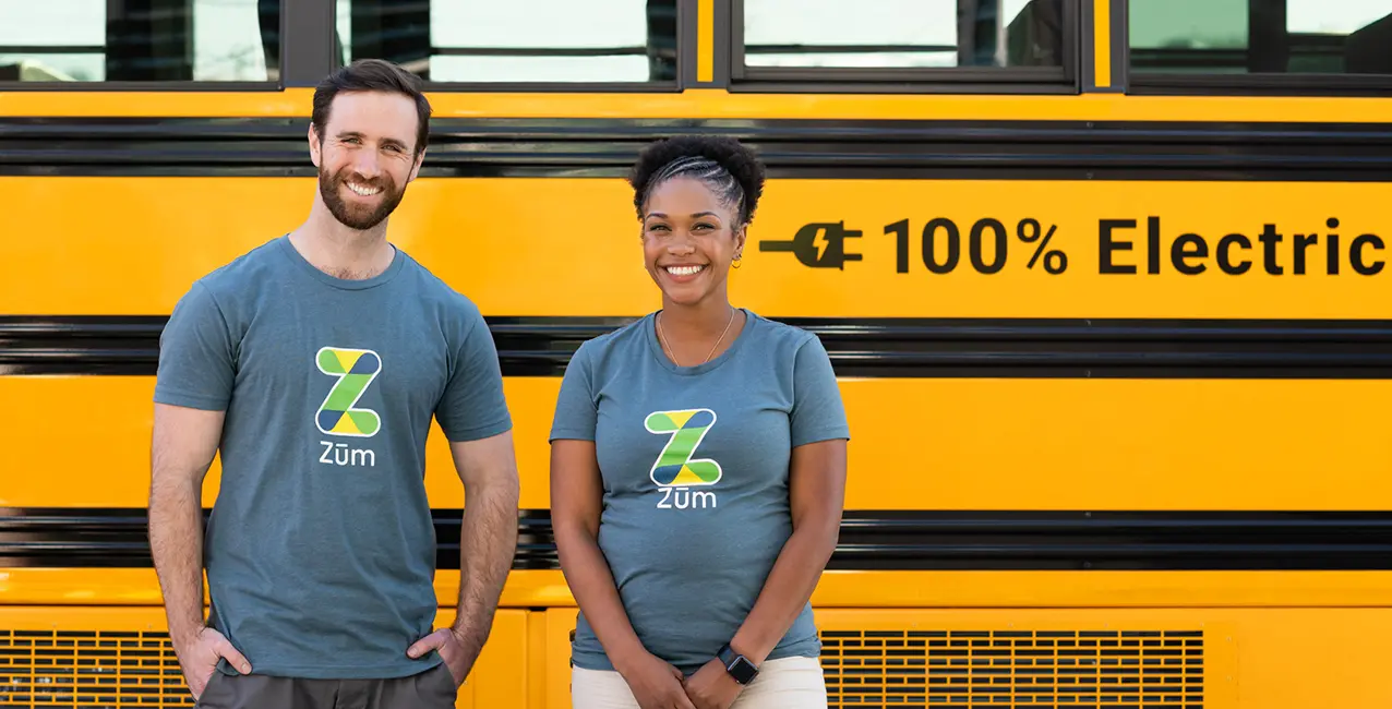Two smiling Zum. team members stand in front of a yellow school bus labeled "100% Electric."