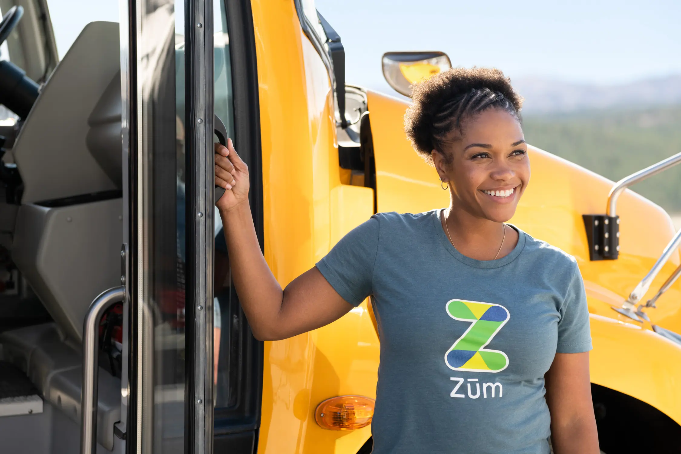 Smiling driver in Zum. shirt stands by a yellow school bus door, ready for passengers.