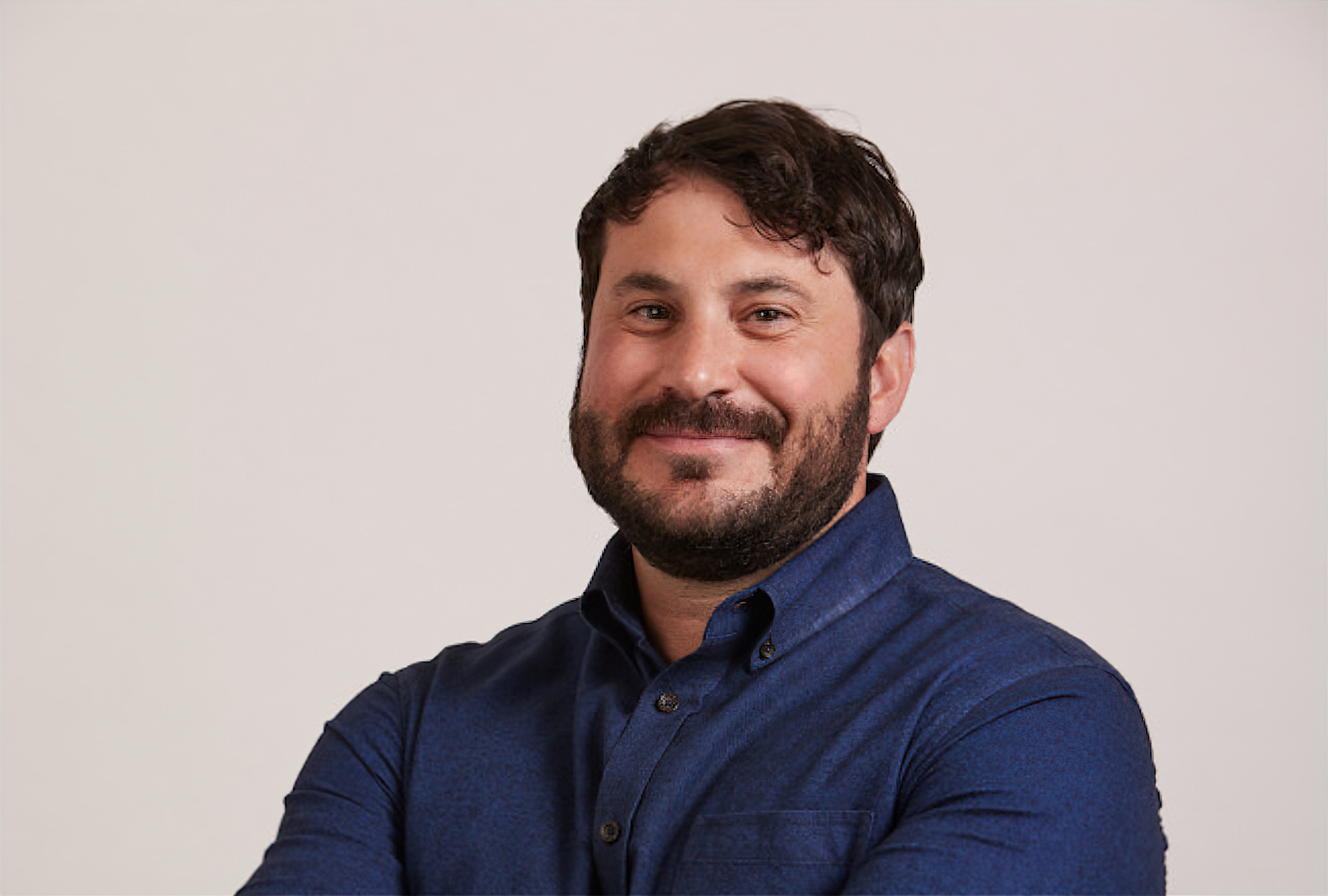 Man with dark hair and beard, wearing a blue shirt, smiling and crossing arms against a light