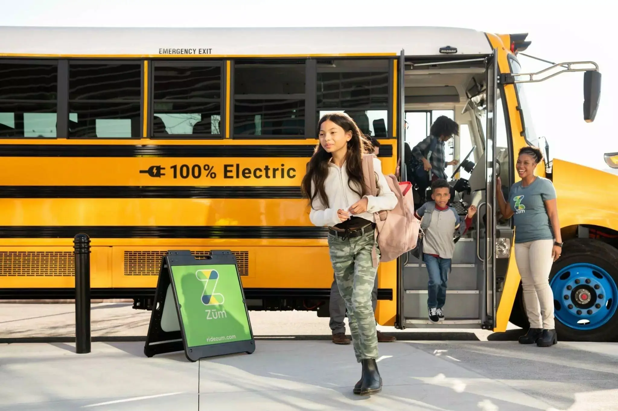 A student exits a Zum electric bus, with parents and a child nearby. A sign reads "100% Electric."