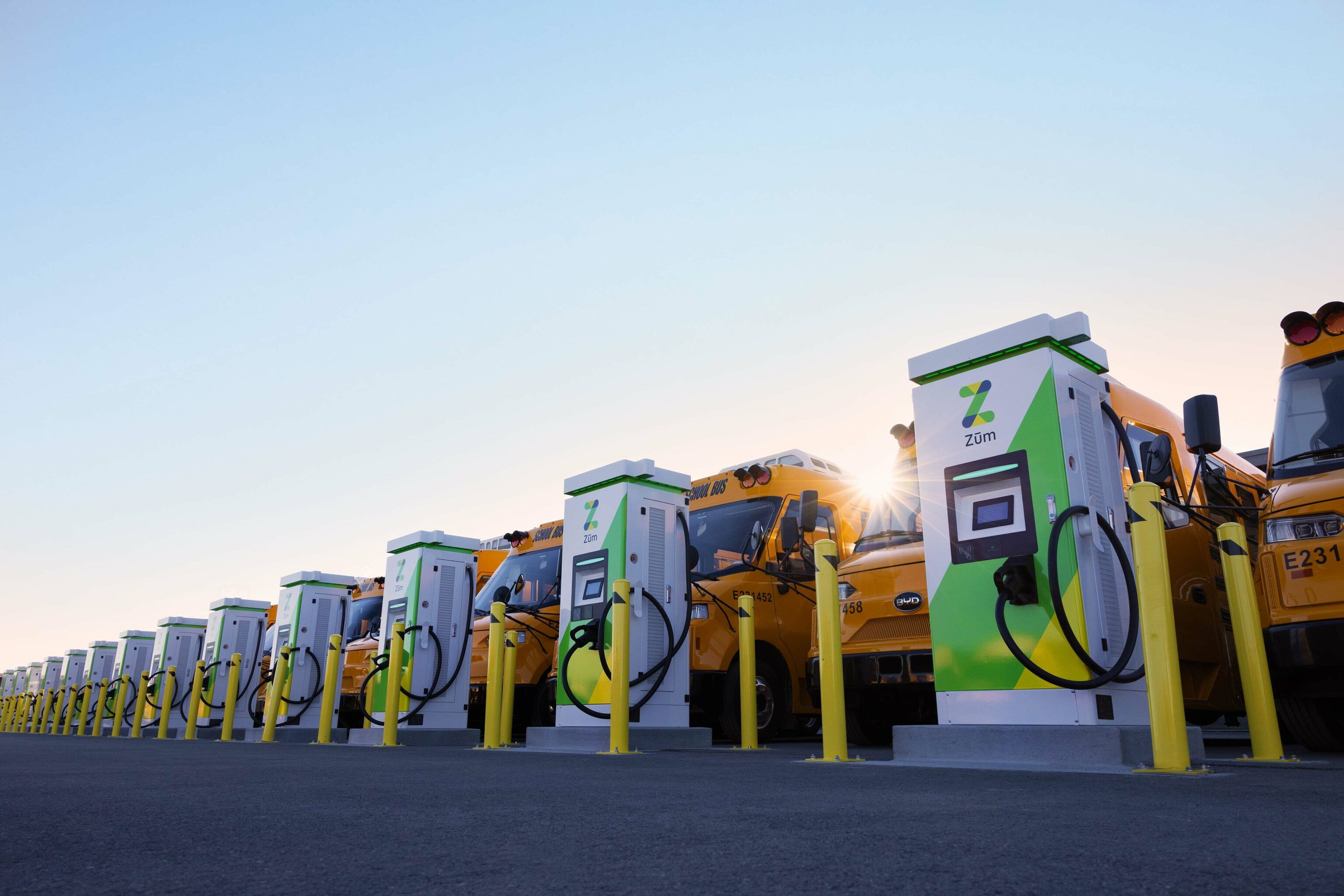 Yellow school buses lined up at EV charging stations in a parking lot, sun shining in the