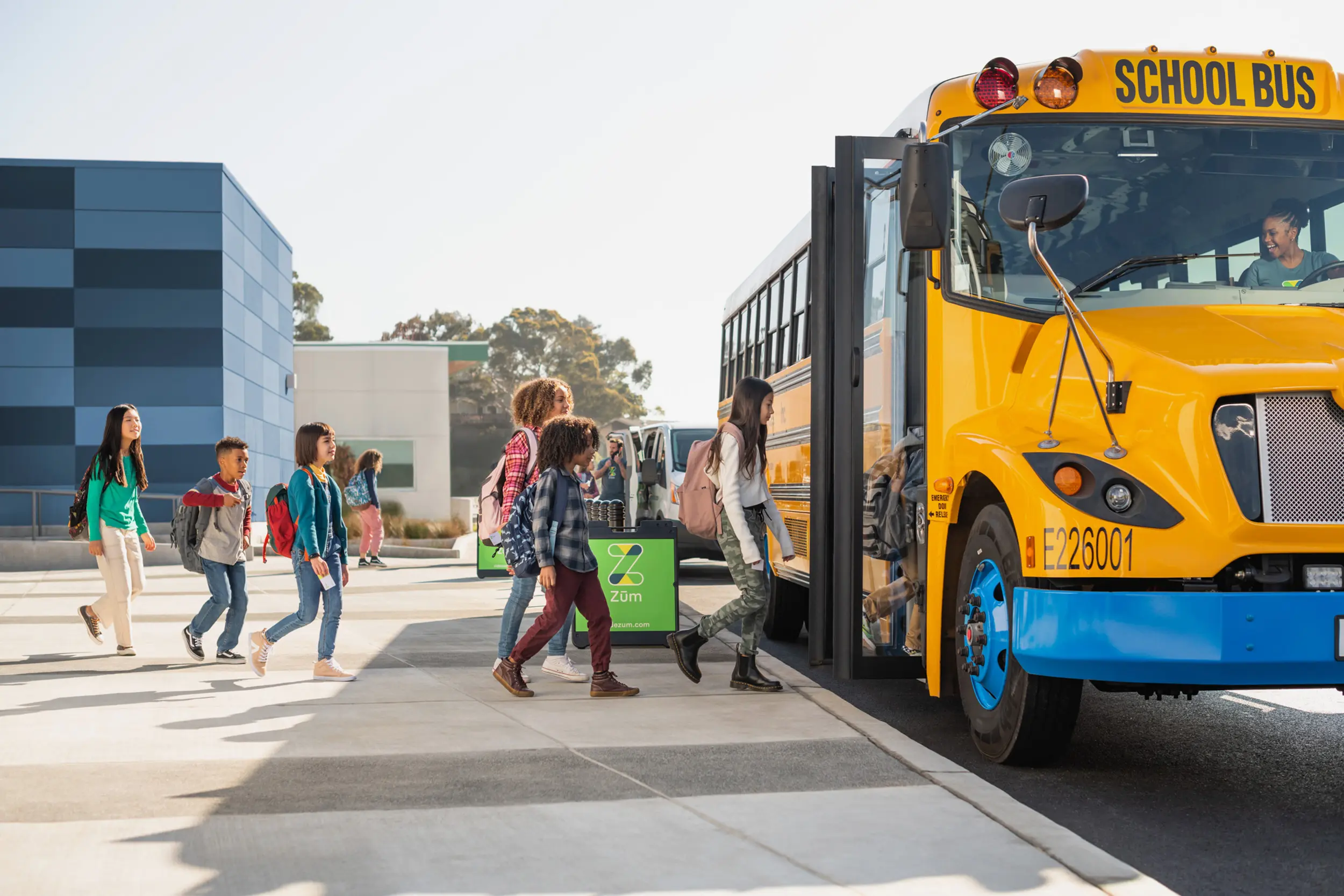 Students are boarding a yellow school bus at a transportation hub on a sunny day.