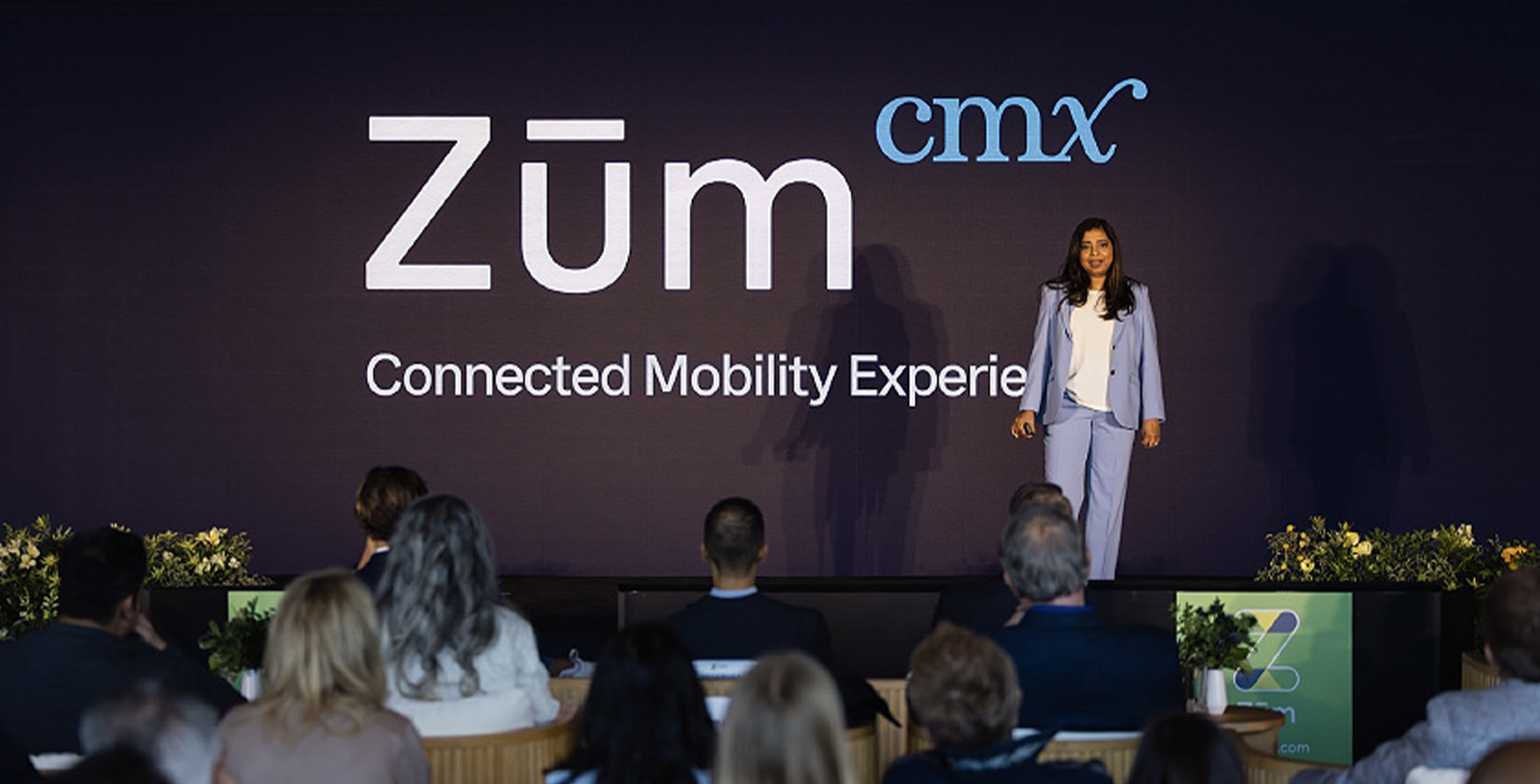 A speaker presents Zum's Connected Mobility Experience at a conference, with an audience visible.