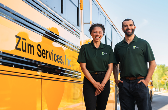 Two smiling professionals in green Zum polos stand beside a yellow school bus, showcasing company
