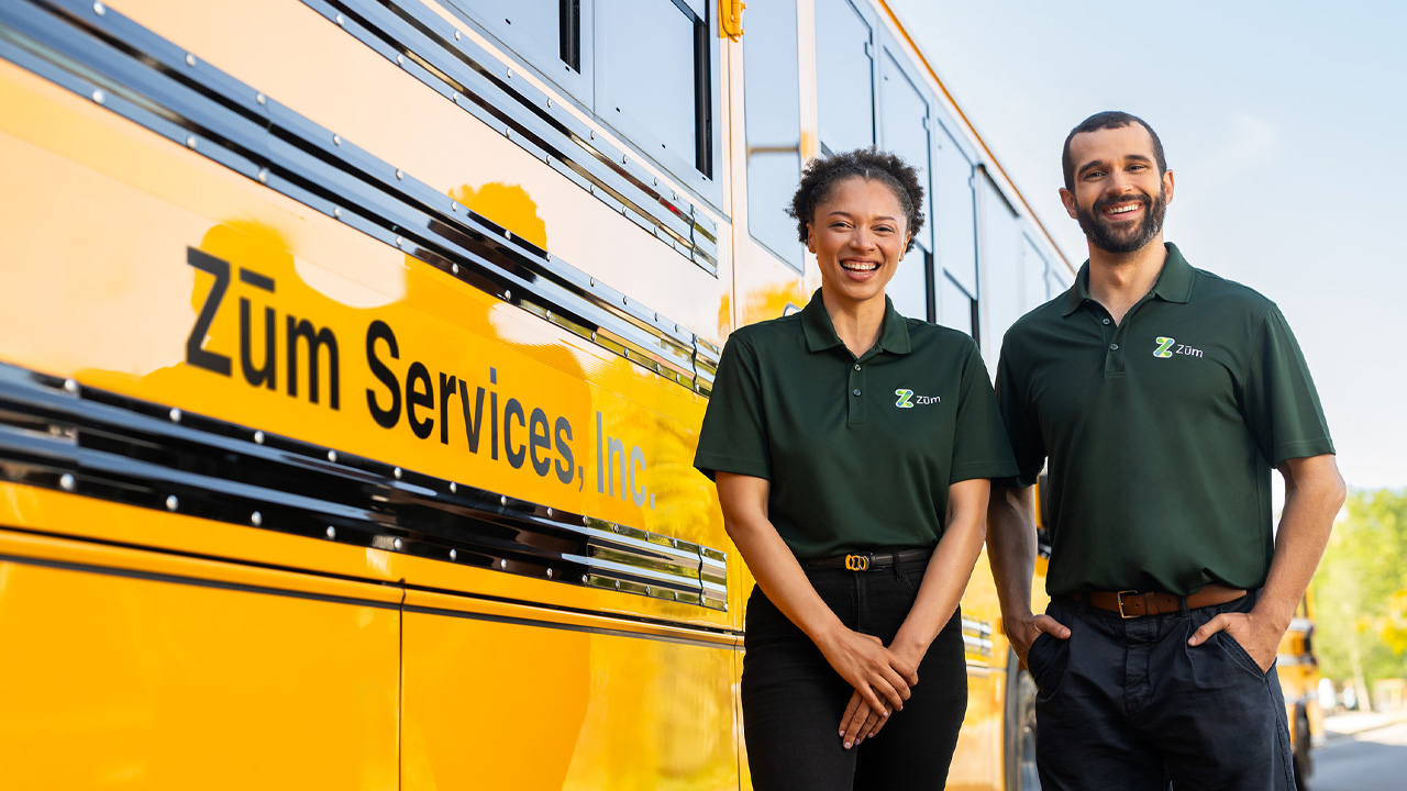 Two Zum employees stand in front of a yellow school bus, smiling and wearing branded green polos.