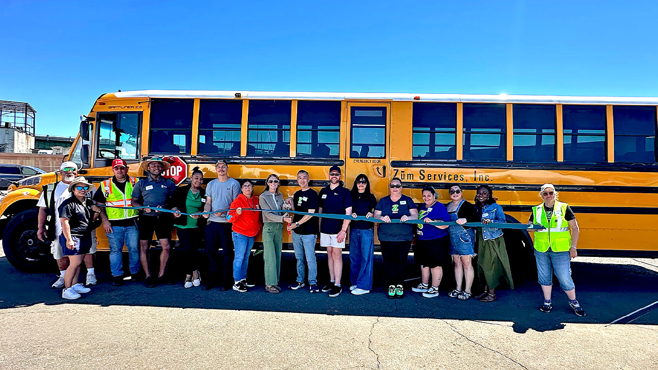 A ribbon-cutting event with employees in front of a yellow Zum school bus.
