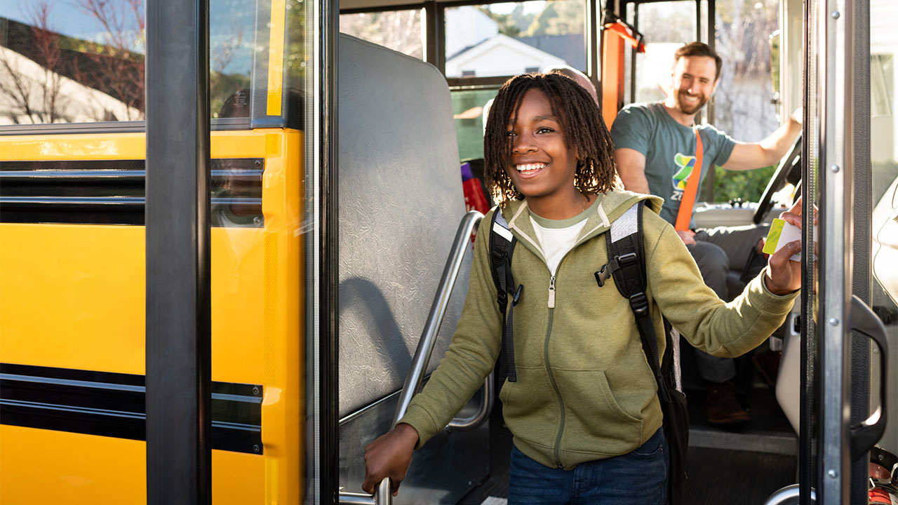 A student with a backpack smiles as they board a school bus, with a driver visible inside.