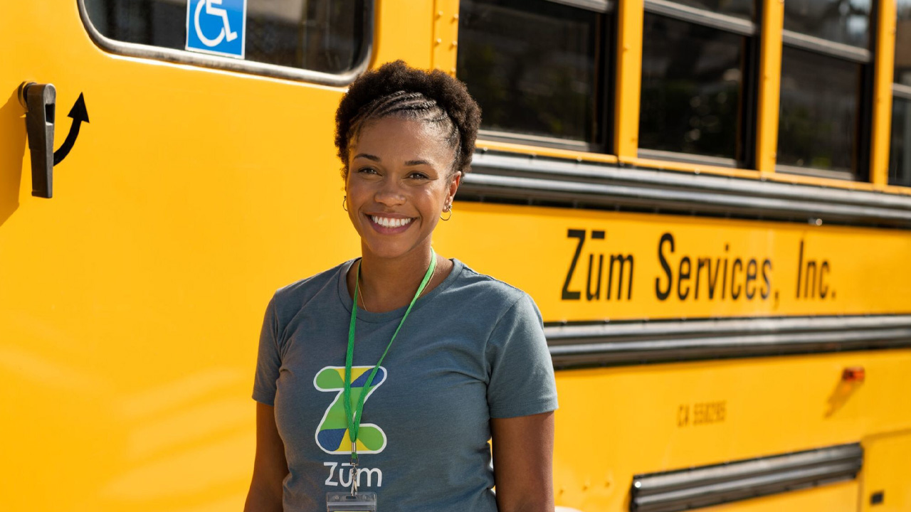 Smiling woman in a Zum. shirt stands in front of a yellow school bus.
