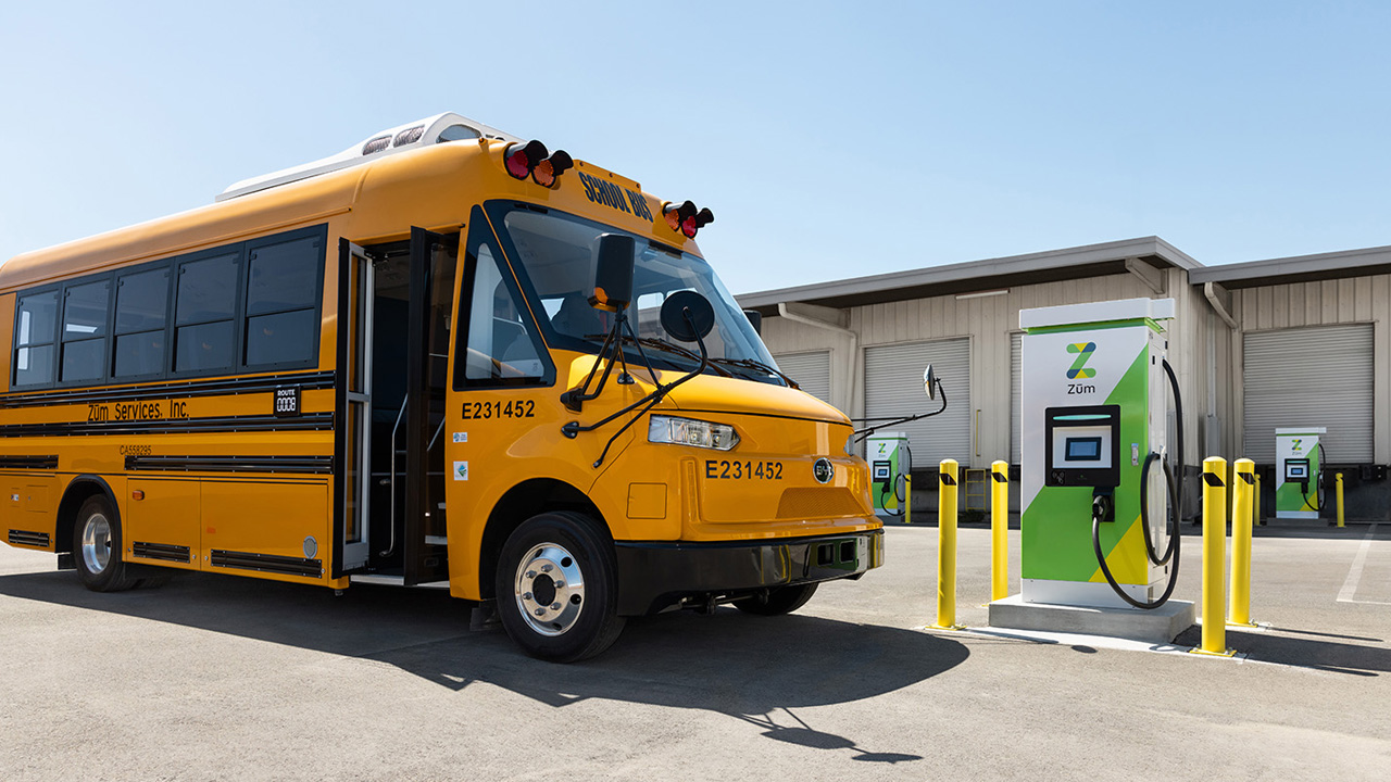 Yellow electric school bus parked next to EV charging station at a bus yard.