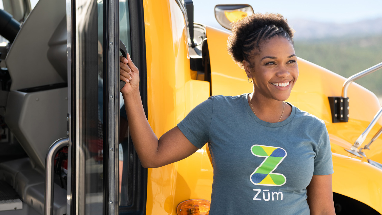A smiling woman in a Zum. shirt stands by a yellow school bus door.