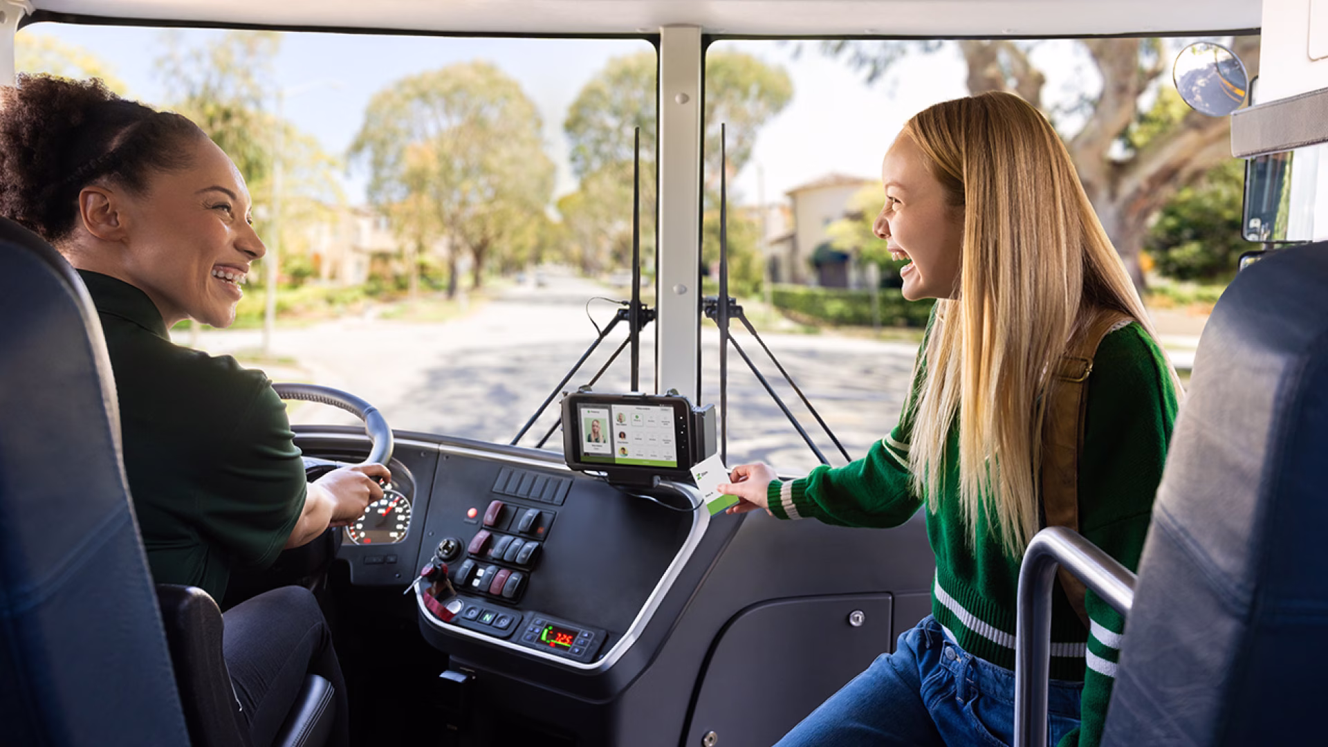 Two women smile inside a bus; one drives while the other engages with a device.