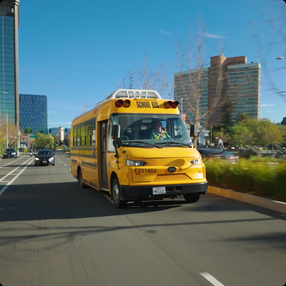 A yellow school bus drives on a city street, with buildings visible in the background.